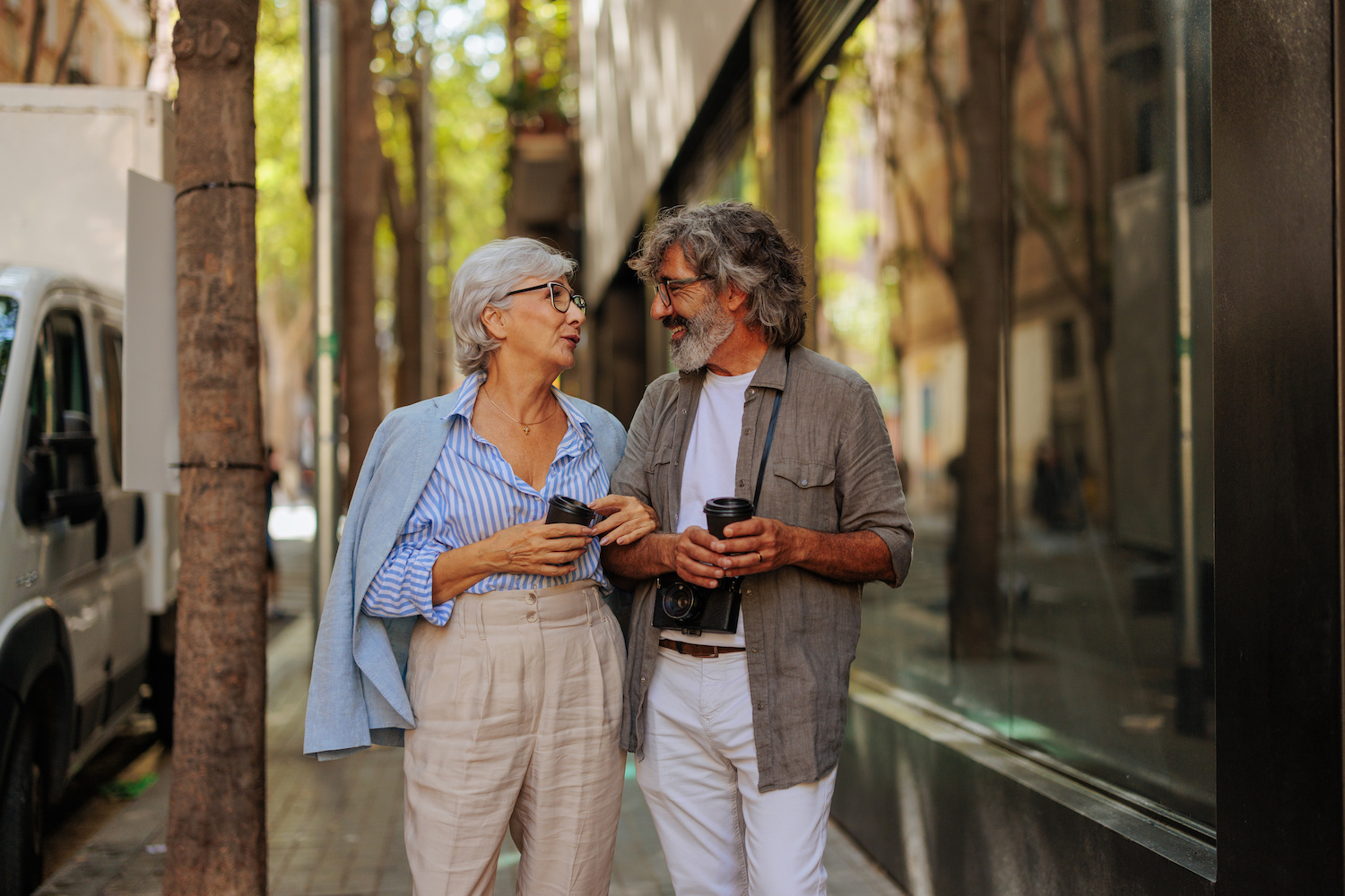 elegant mature couple holding coffees walking down city sidewalk